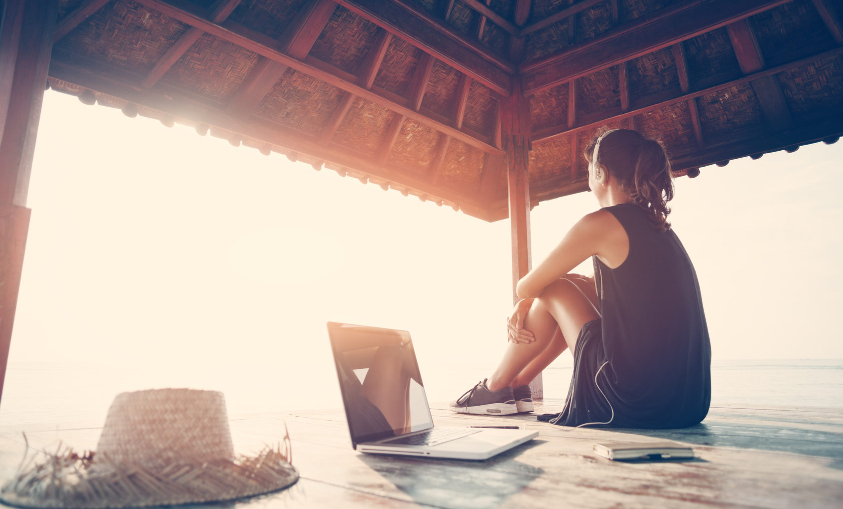 Woman freelancer listening music at sunrise near ocean in shadow shelter. Intentional sun glare and vintage color